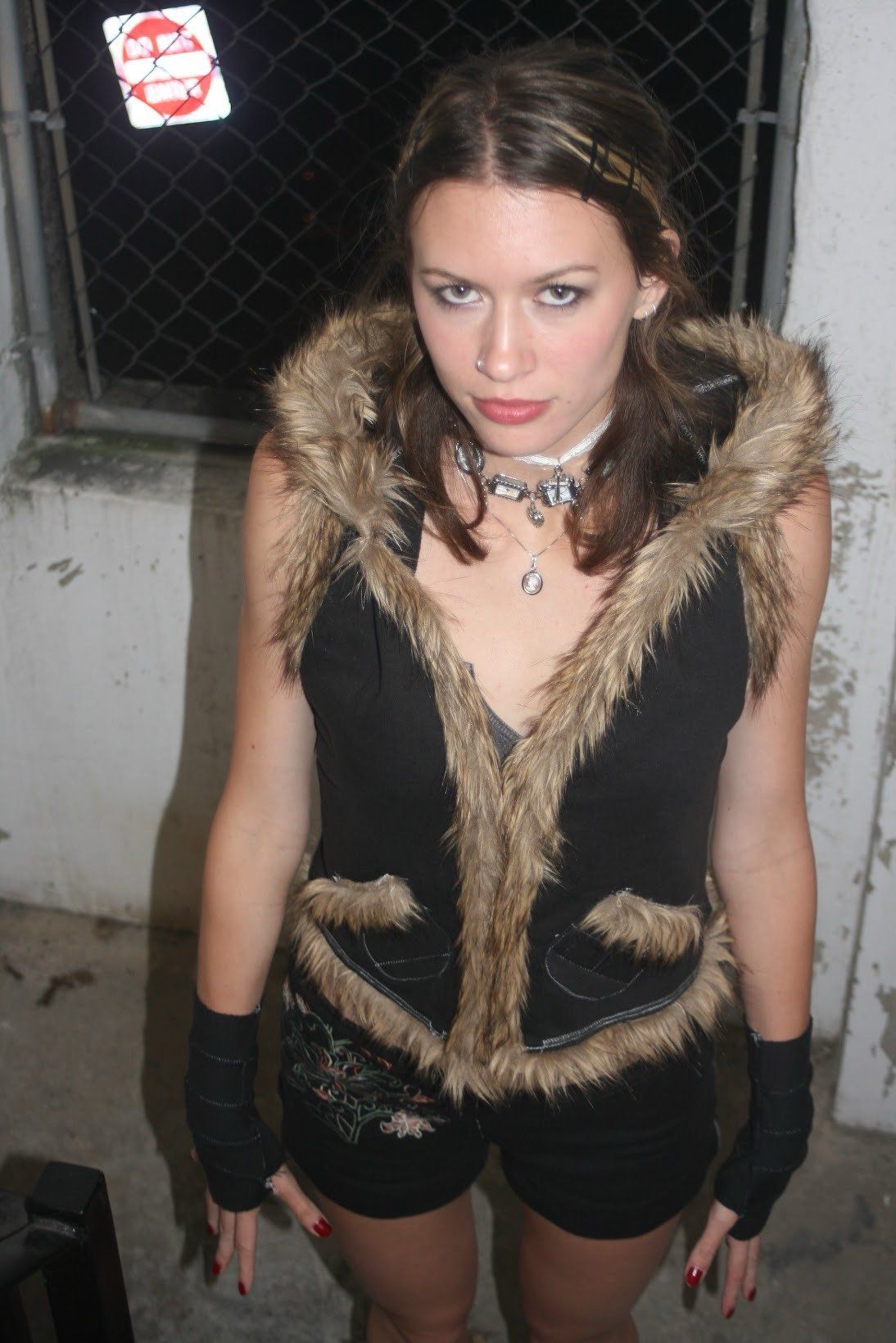 Front view of the black hooded vest with fur trim along the hood and front edges, worn with embroidered black shorts and black fingerless gloves, photographed at night.
