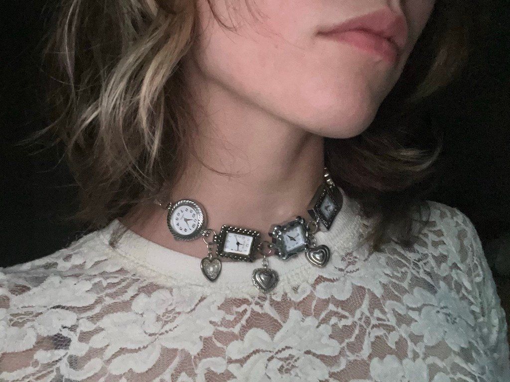 Choker of linked watch faces with heart charms, worn over a white lace top.