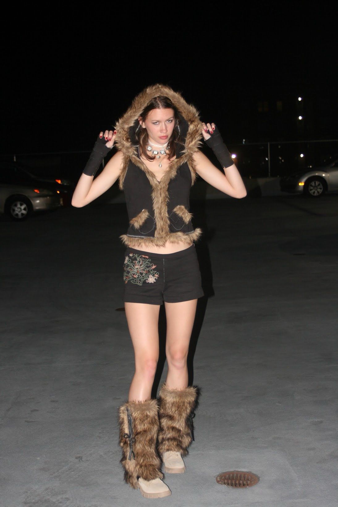 Full-length view of the model holding up the fur-trimmed hood, wearing the vest, embroidered black shorts, and furry leg warmers in a parking lot at night.