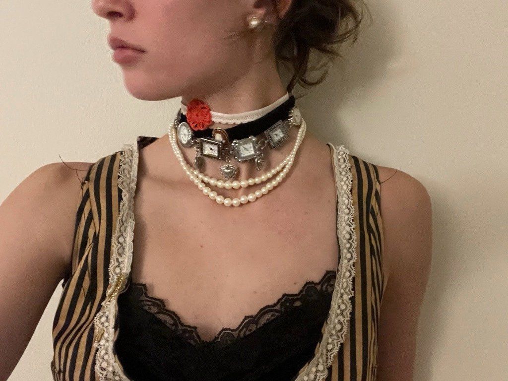 Watch-face choker with a red flower, layered over pearl strands and worn with a striped vest trimmed in lace.