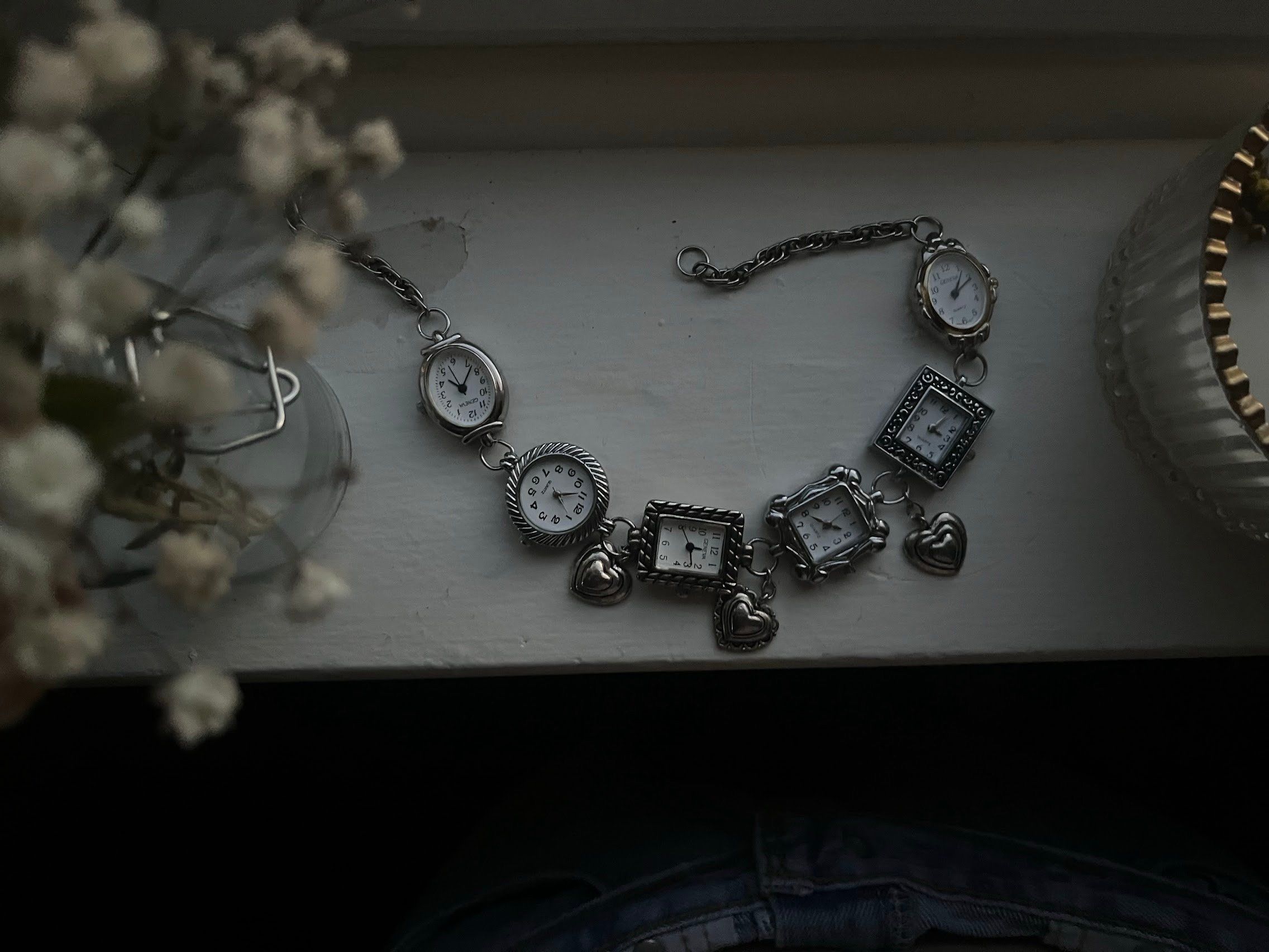 Chain of linked watch faces with heart charms, laid flat on a white windowsill beside baby's breath.