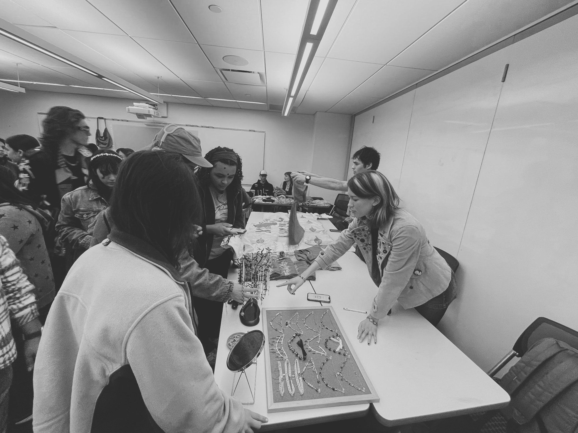 Shoppers crowd a classroom pop-up shop, browsing necklaces on a display board and jewelry tree, in black and white.