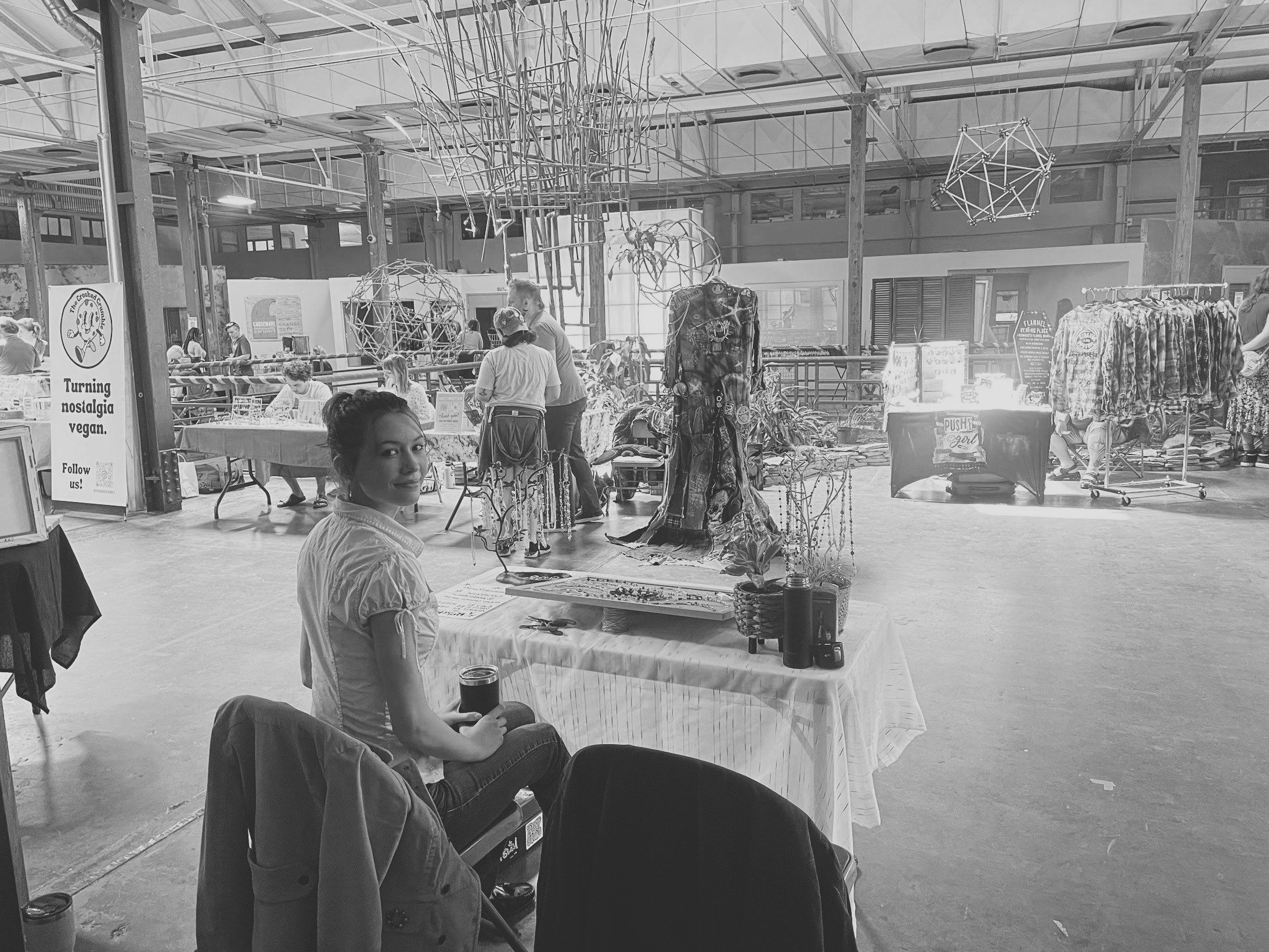 A vendor seated beside a jewelry table at an indoor market in a large warehouse hall, in black and white.