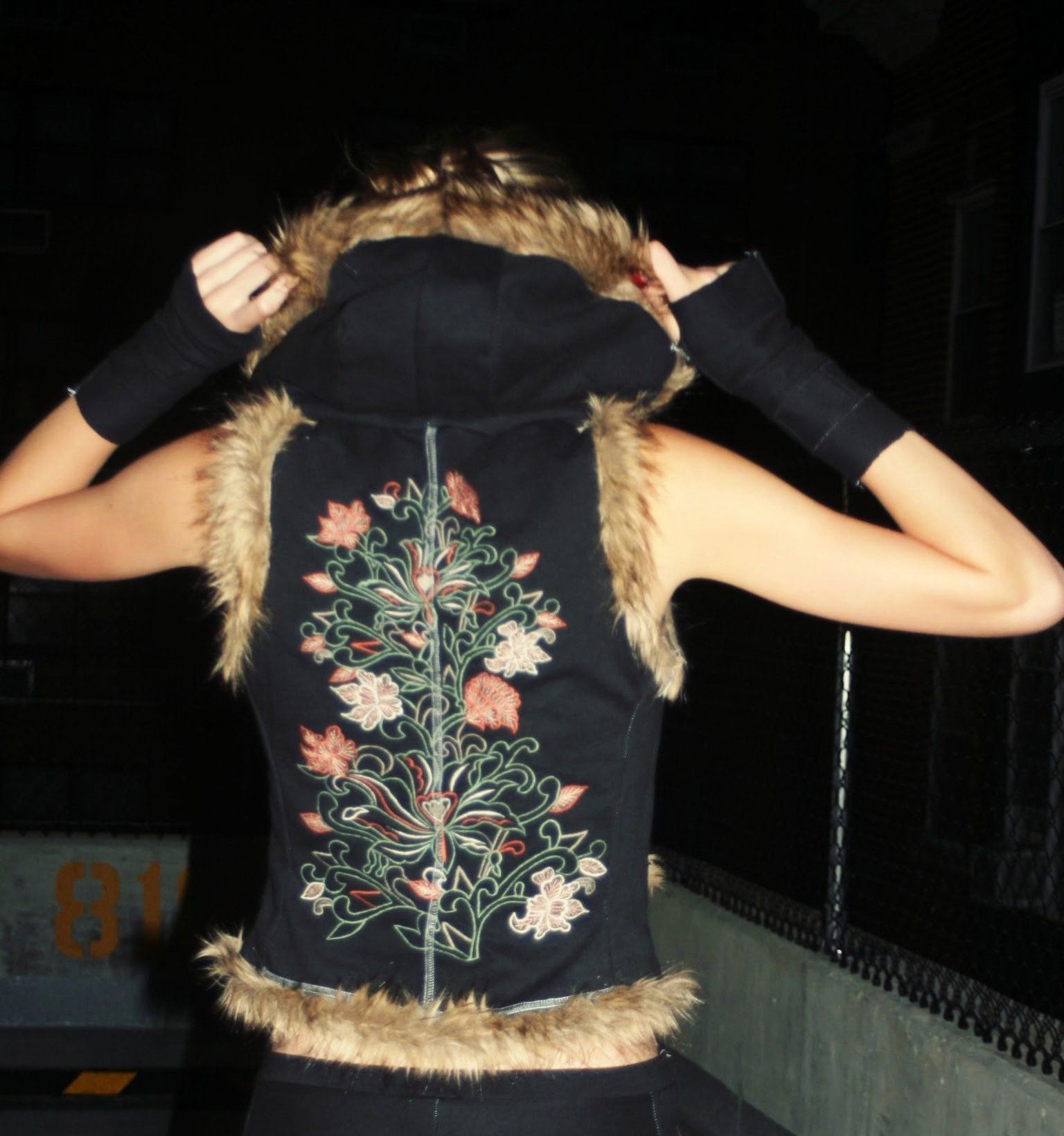 Back view of a model pulling up the hood of a black vest with fur trim, its back covered by a tall floral embroidery panel, at night by a chain-link fence.