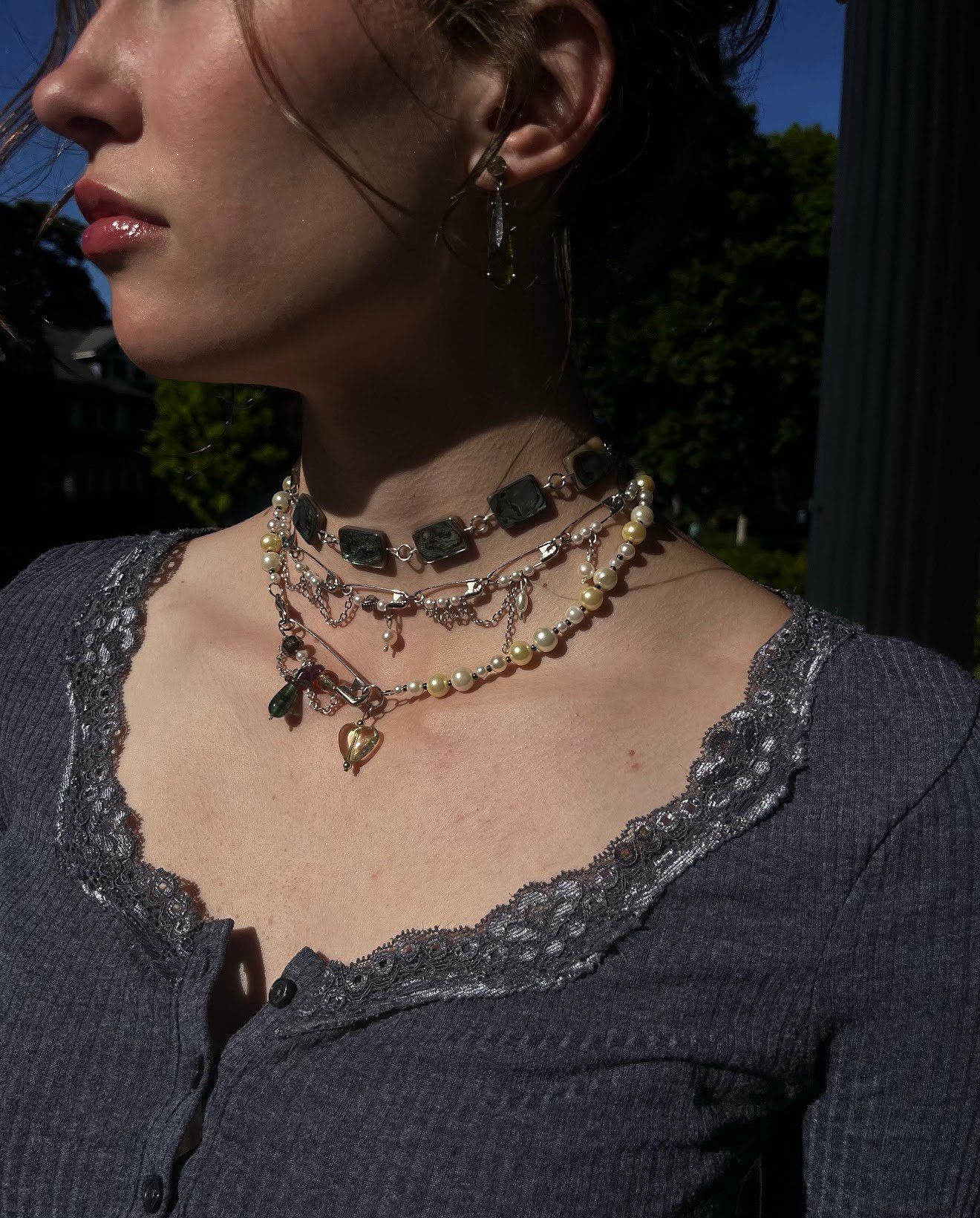 Layered necklaces with square gray-green links, pearl strands, safety pins, and a glass heart charm, worn with a gray lace-trimmed top, shown in profile outdoors.