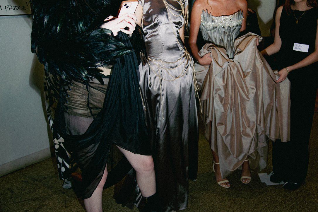 Backstage, three models in Look 2's feather jacket and chiffon skirt, Look 3's beaded silver satin dress, and Look 4's feathered gown, shown from the shoulders down.