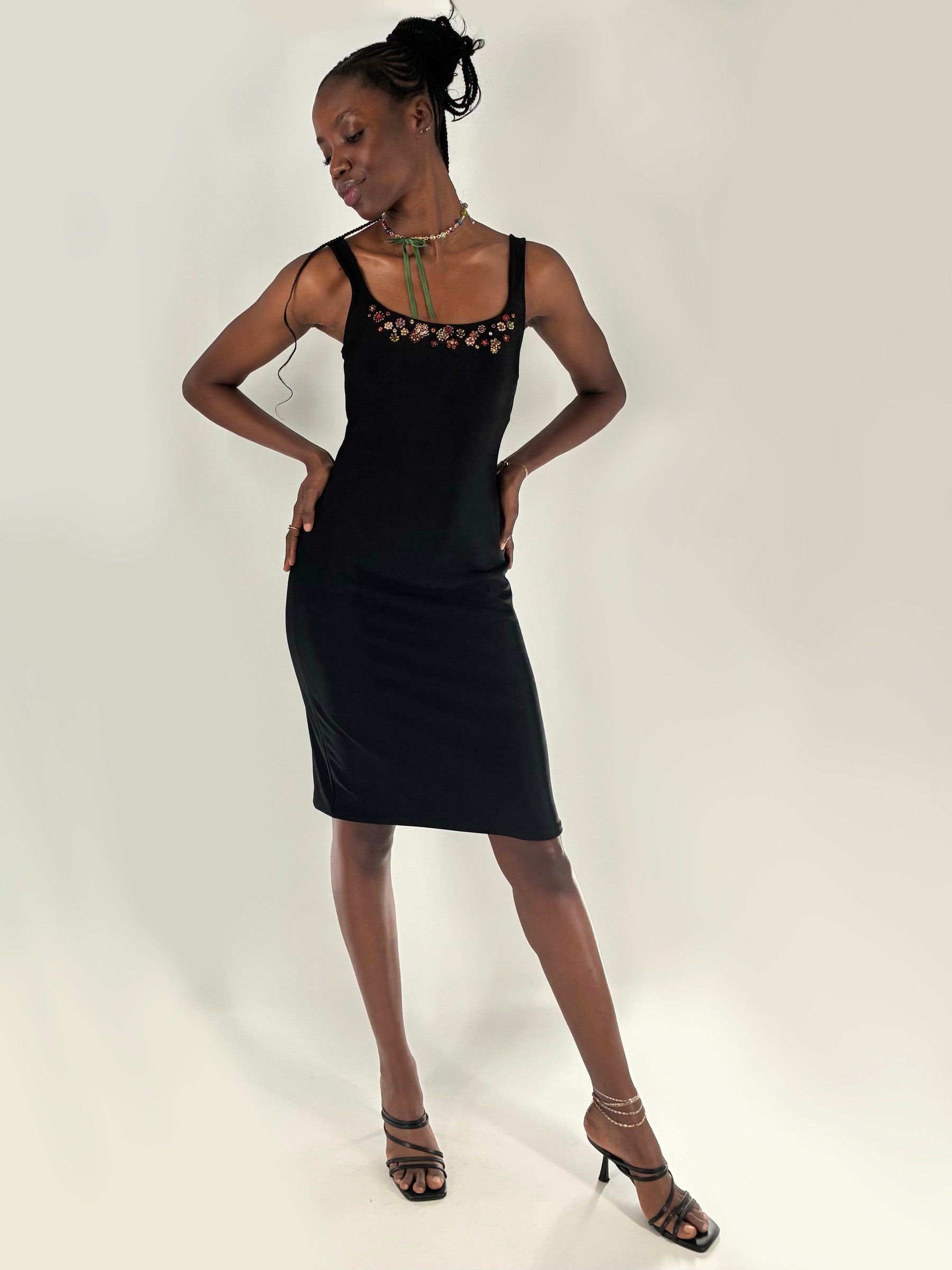 Model in a fitted black knee-length knit dress with a glass-bead floral neckline and a beaded choker tied with green ribbon, hands on hips, in the studio.