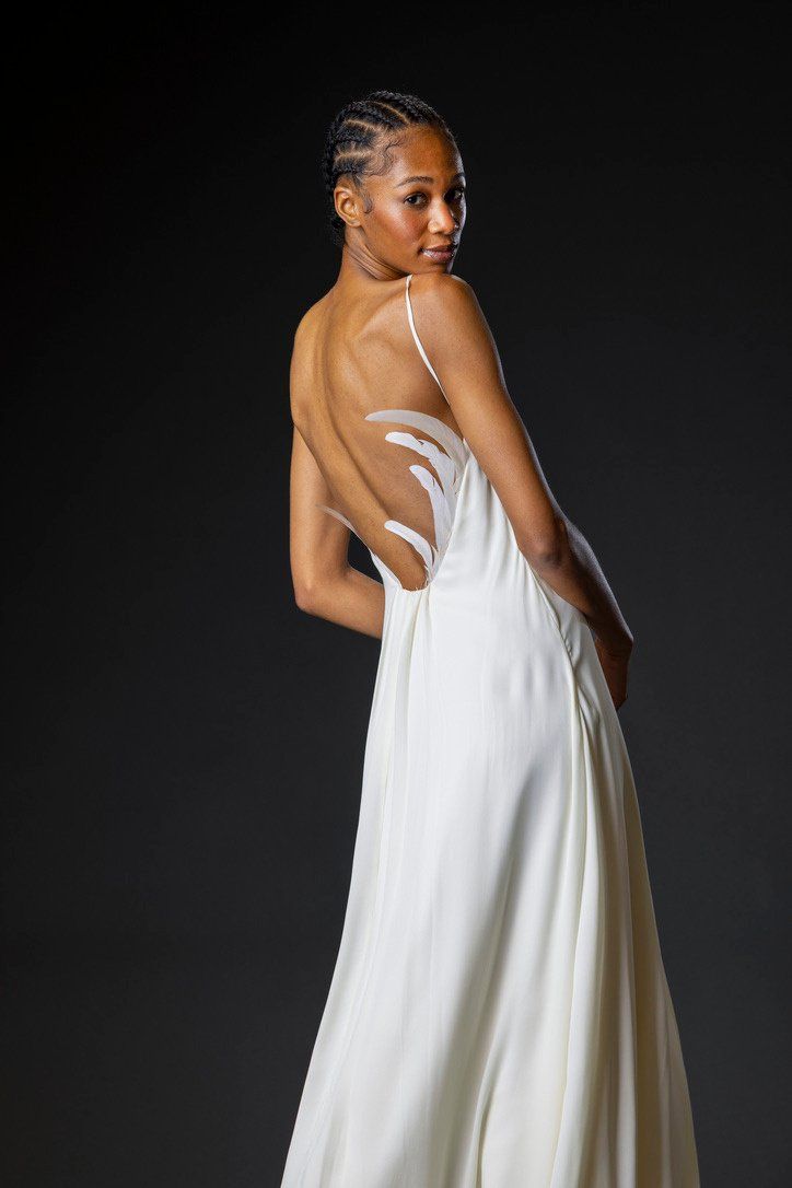 Back view of the ivory silk georgette gown, showing the open back edged with white rooster tail feathers, on a dark studio backdrop.