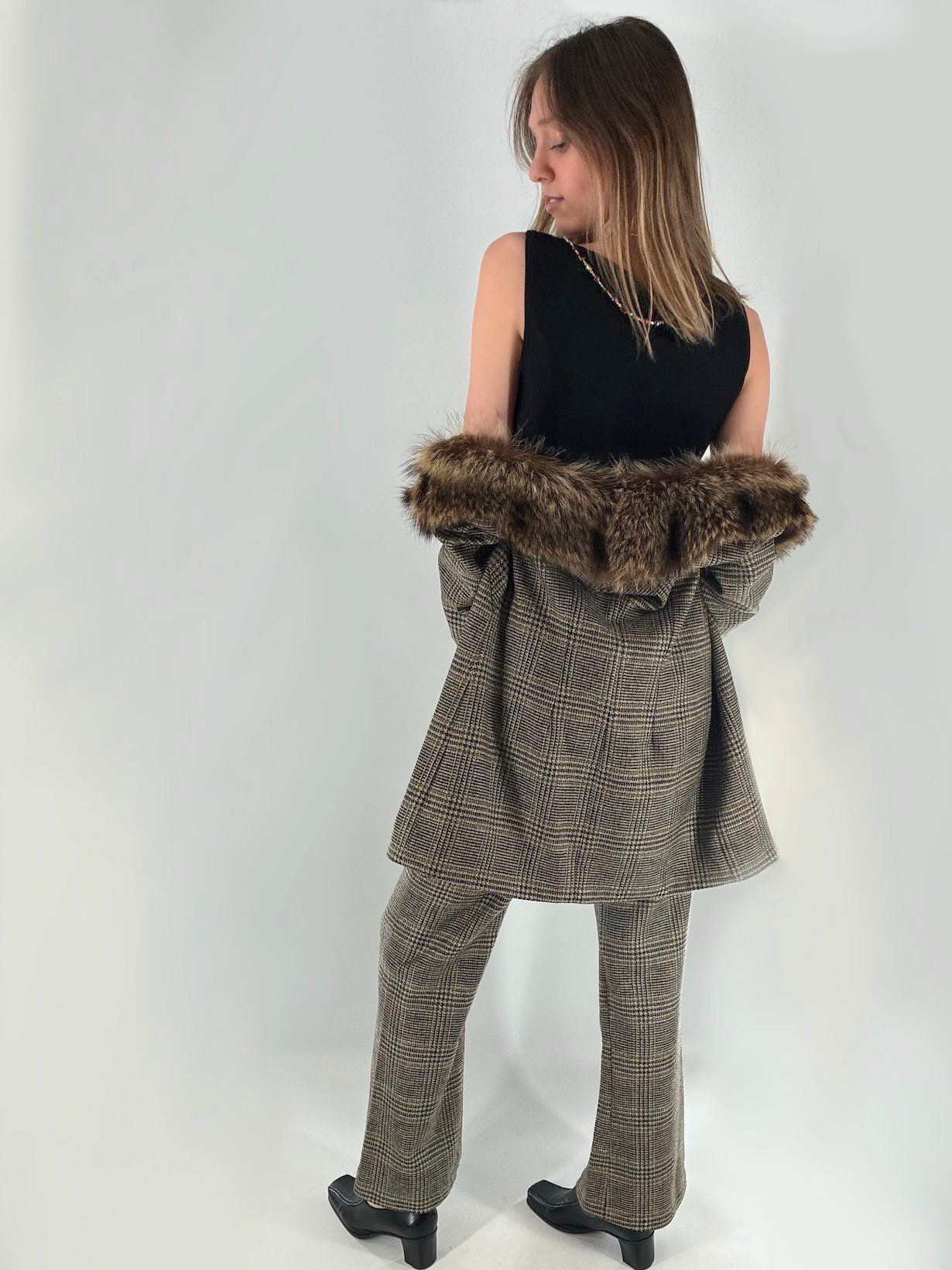 Back view of a brown plaid wool suit with flared trousers, the long jacket slipped off the shoulders to show its fur collar over a sleeveless black knit top.