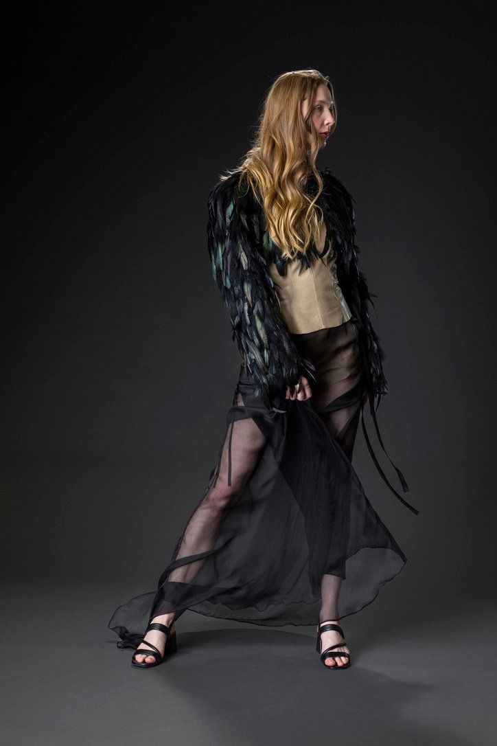Full-length view of Look 2: a cropped black rooster tail feather jacket, tan leather corset, and sheer black silk chiffon overskirt with a train.