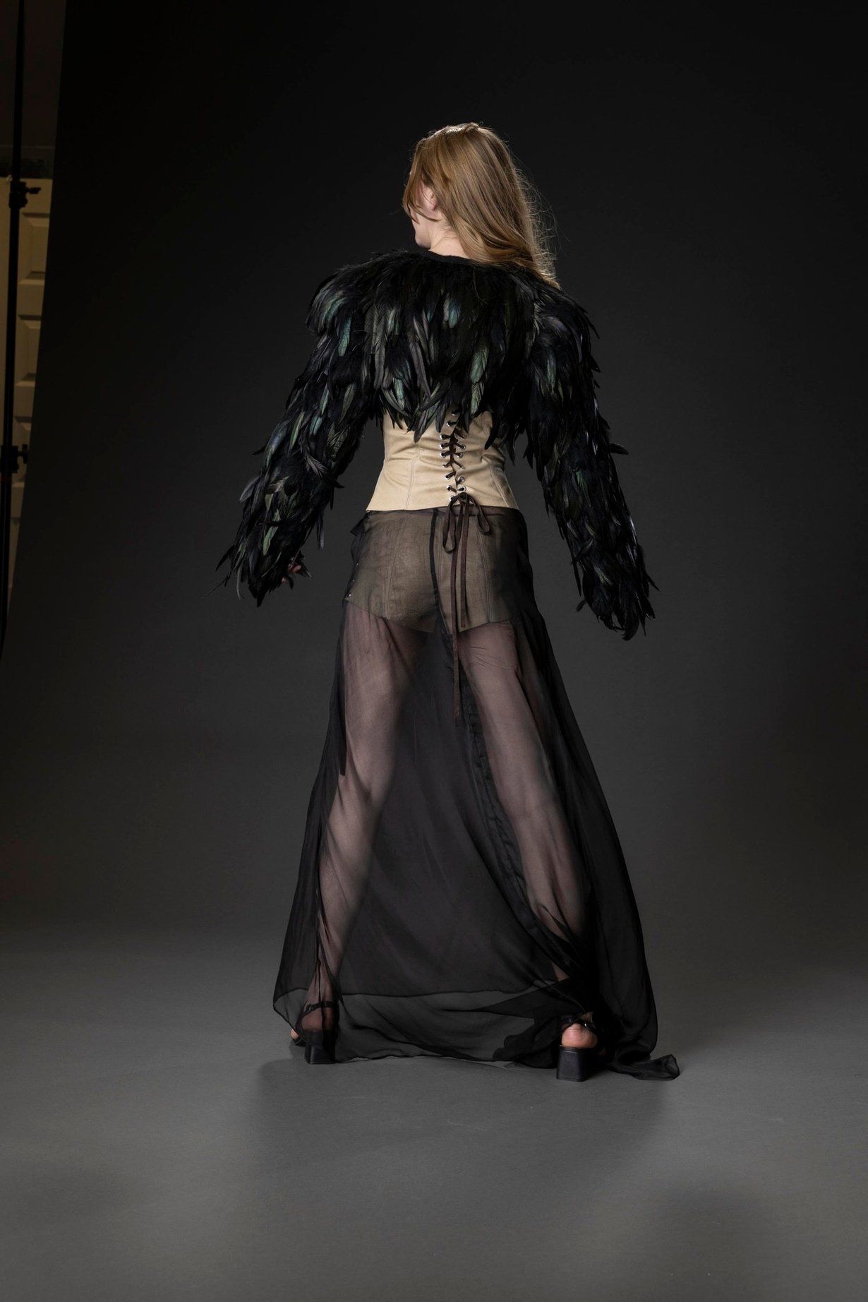 Back view of Look 2 showing the cropped feather jacket, the corset's lace-up back, and the sheer chiffon overskirt.