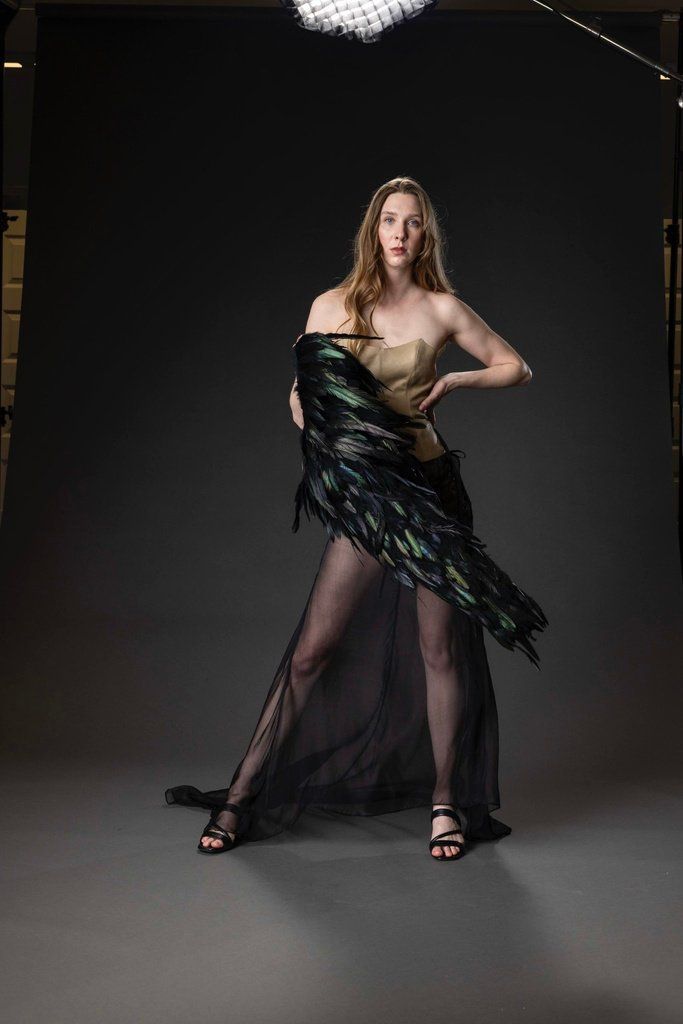 Front view of Look 2 with the feather jacket slipped off one shoulder, showing the tan leather corset and sheer overskirt.