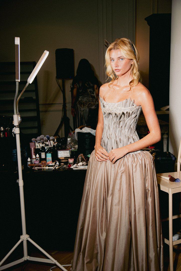 Backstage, the model in the strapless Look 4 gown with a gray feather bodice, jeweled neckline, and full gathered taupe skirt, beside a makeup station.