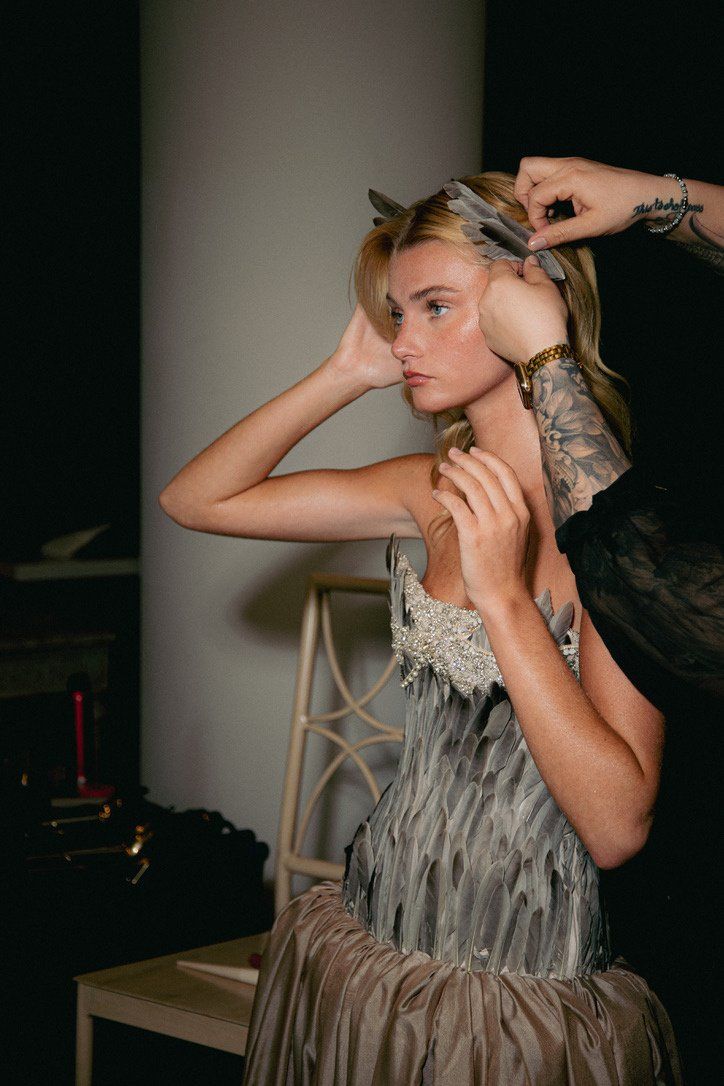 Backstage close-up of the Look 4 model having a feather hairpiece adjusted, showing the jeweled trim at the neckline.