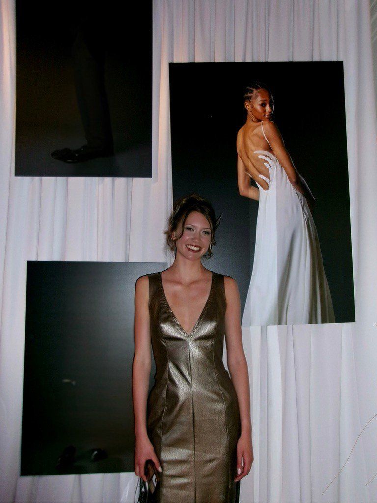 The model smiling in the metallic faux leather dress with a plunging V neckline, in front of a wall hung with photos from the collection.