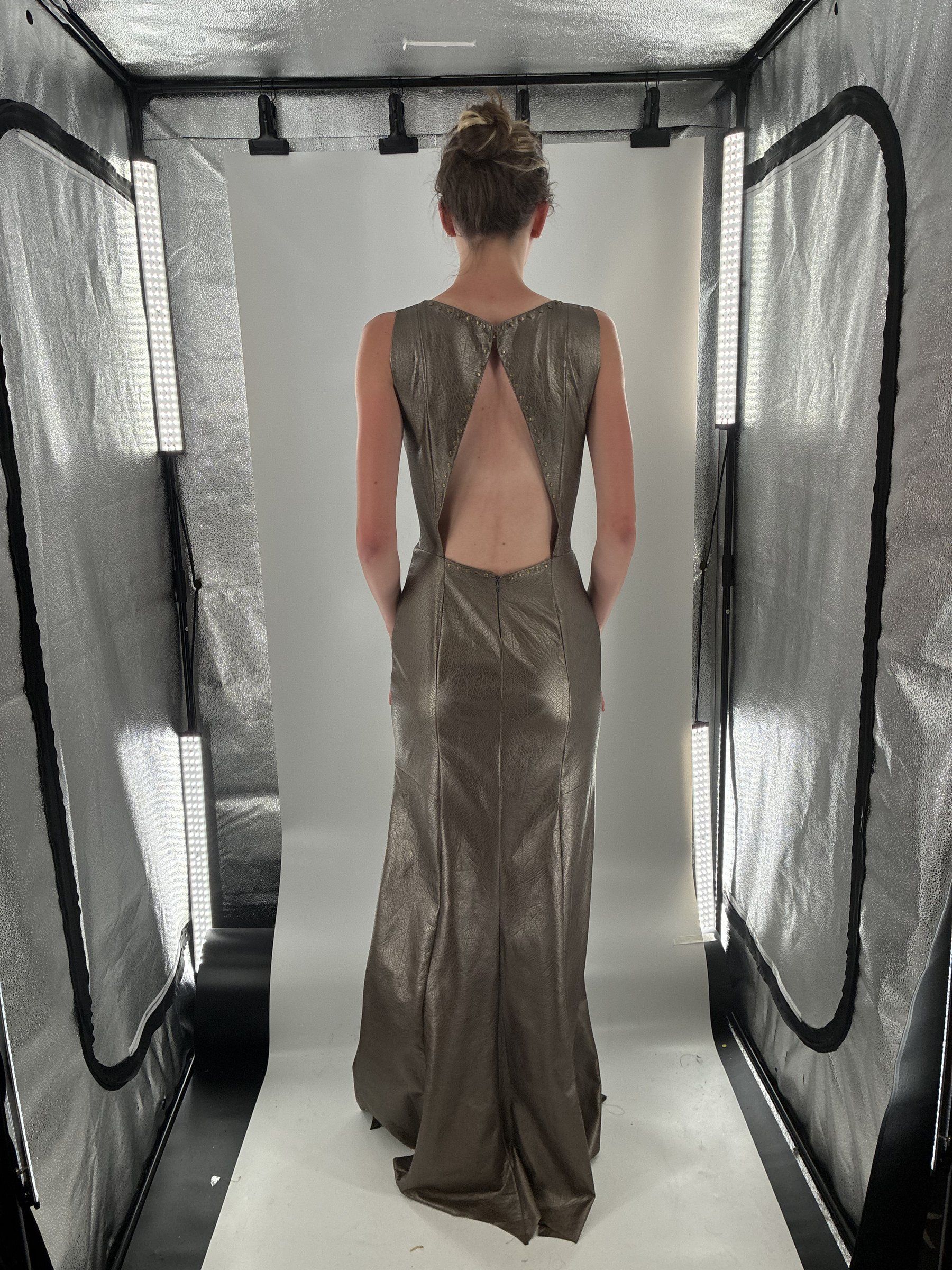 Back view of the full-length metallic faux leather dress in a lit photo tent, showing the back cutout edged with bronze studs.